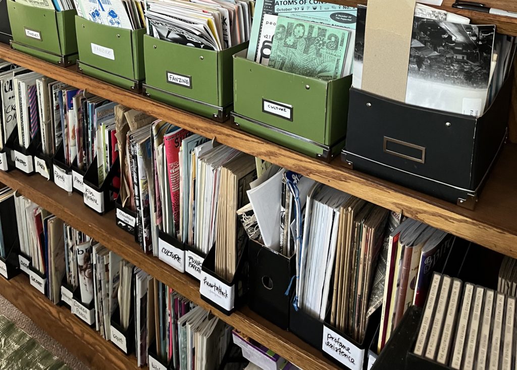 A photograph of wooden shelves holding storage cases containing various zines. 